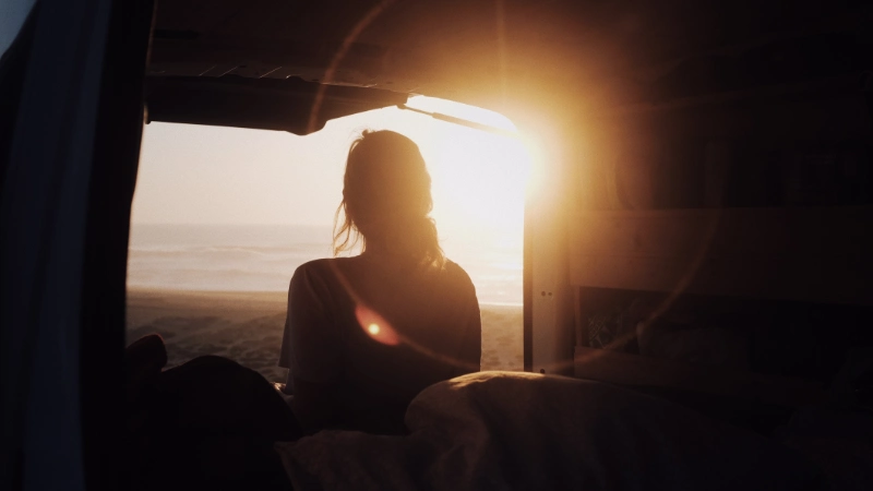 Eine Junge Frau sitzt im Minicamper und schaut sich den Sonnenuntergang an