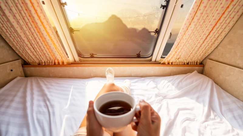 Eine Frau liegt mit einer Tasse Kaffee in Ihrem Minicamper und schaut sich den Sonnenaufgang an